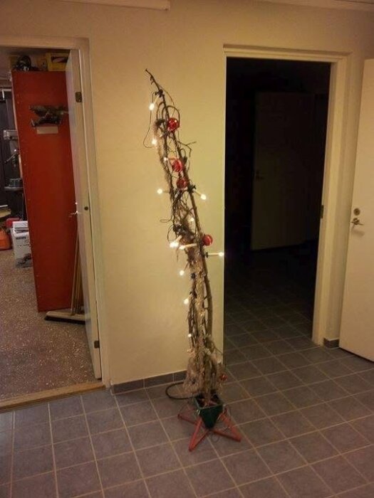 En smal julgran med röda kulor och ljus i en hall med två dörrar.