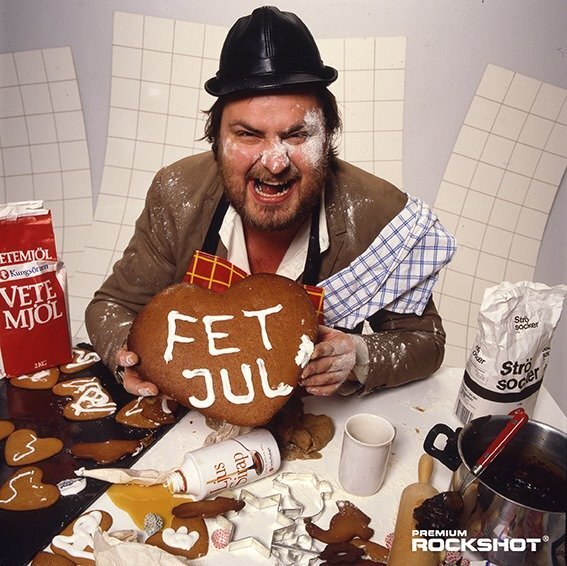 En man i hatt grimaserar och håller ett pepparkakshjärta med texten "FET JUL" omgiven av bakkaos med mjöl och socker.