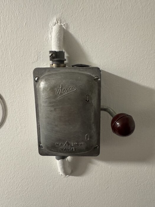 Metallisk elbox med texten "Wesco", monterad på en vägg, med en ansluten kabel och en röd, rund spak till höger.