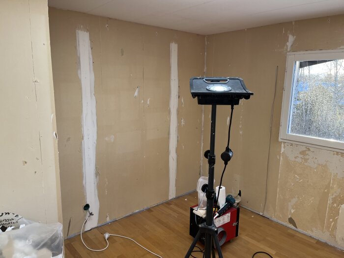 Rum under renovering med avskalade väggar och spacklade skarvar, ett fönster och en arbetslampa på stativ. Verktyg och material på golvet.