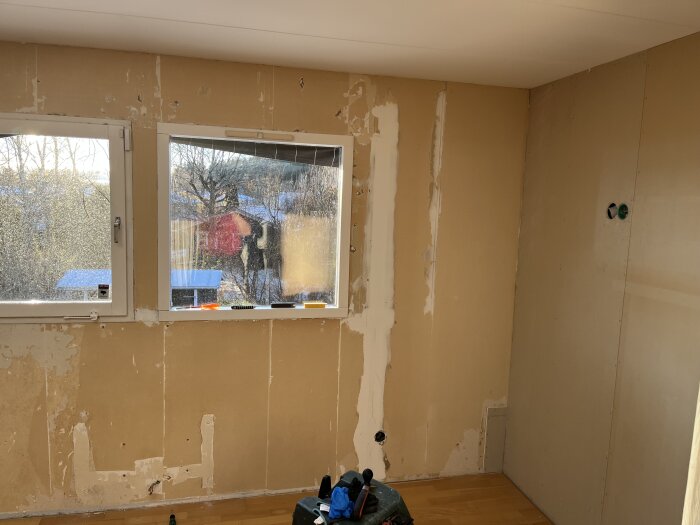 Renovering av rum med borttagna tapeter och spacklade väggar, två fönster och verktyg synliga inuti rummet.