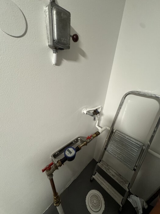 Källarvägg med vattenrör, manometer och stege. Enhet i hörnet med anslutningar, avlopp och fjärrvärmecentral i närheten.