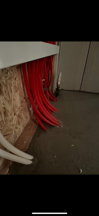 Röda och vita golvvärmeslangar installerade i ett vattenskåp, med slangarna som kommer upp från golvet nära väggen.