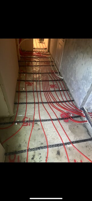 Golvvärmerör installerade i ett hus, synliga på betonggolvet med markerade linjer; arbetet verkar ofärdigt och kräver justeringar.