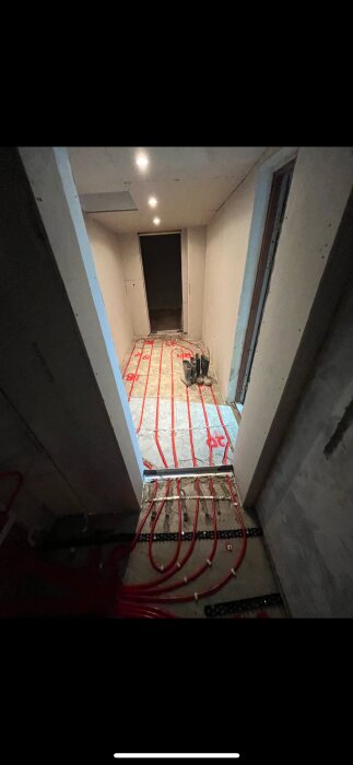 Byggarbetsplats med golvvärmerör monterade på cementgolv, ljussatta av spotlights i taket; stövlar står i hörnet.