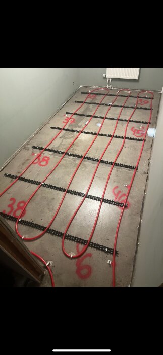 Golvvärmesystem med röda rör på betonggolv, numrerade markeringar synliga, omgivning med vita väggar.
