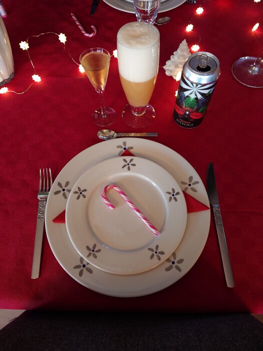 Juldukning med röd duk, ljusslinga, godiskäpp på tallrik, snaps, ölglas och burk med "Juleljus" på en bordsscen.