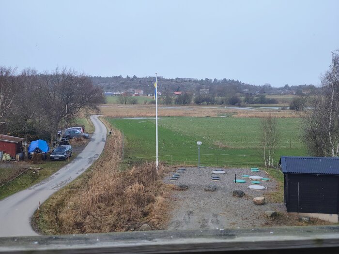 Vinterlandskap med lätt snötäcke på en landsväg och fält, omgiven av träd och flaggstång.
