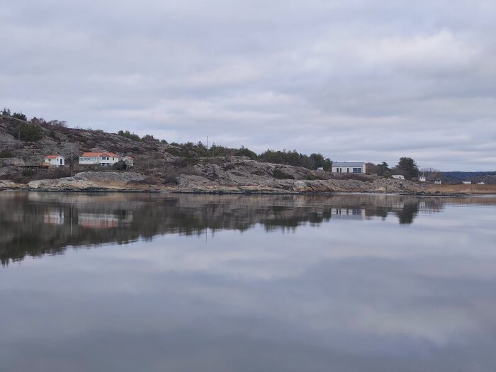 Kustlandskap med klippor och hus vid vatten, molnig himmel speglas i den stilla ytan.
