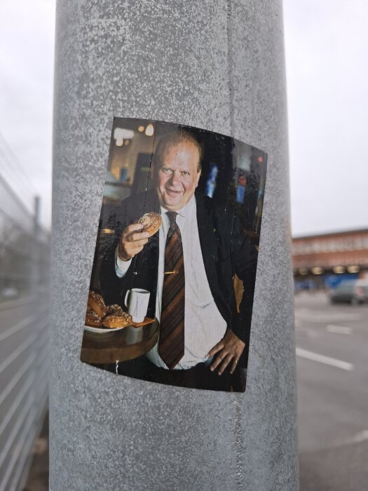 Fotografi av en man i kostym som håller en bulle. Bilden är fastsatt på en metallstolpe i ett utomhusområde.