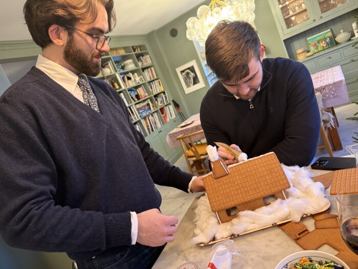 Två personer bygger ett pepparkakshus på julafton i ett kök; bordet är täckt med bomull och pepparkaksdelar.