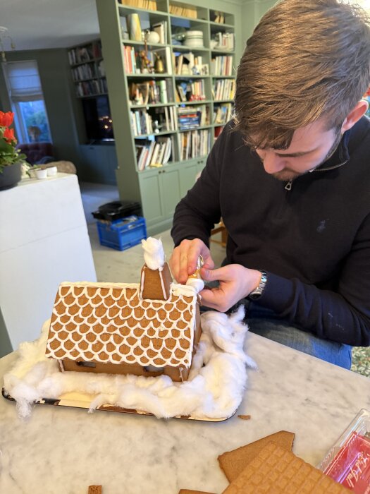 En person dekorerar ett pepparkakshus med glasyr på ett bord, omgiven av bomull som föreställer snö, framför en bokhylla.
