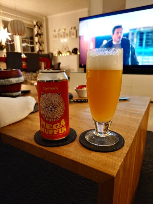 Burk med Stigbergets öl "Mega Muffin" och ett glas öl står på ett bord i ett vardagsrum med en tänd TV i bakgrunden.