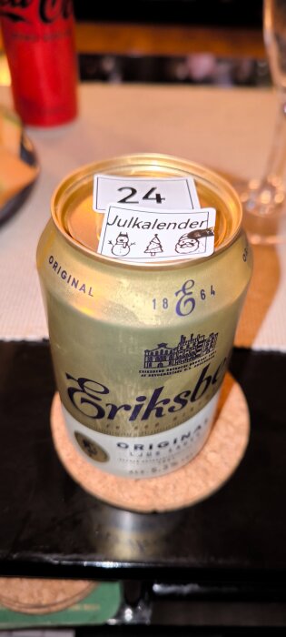En ölburk märkt med en julkalenderetikett med numret 24, snögubbe, julgran och tomte, placerad på ett underlägg.