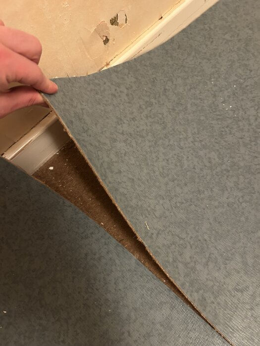 En hand lyfter en del av en grå golvmatta för att visa undersidan och en del av golvet i ett rum under renovering.