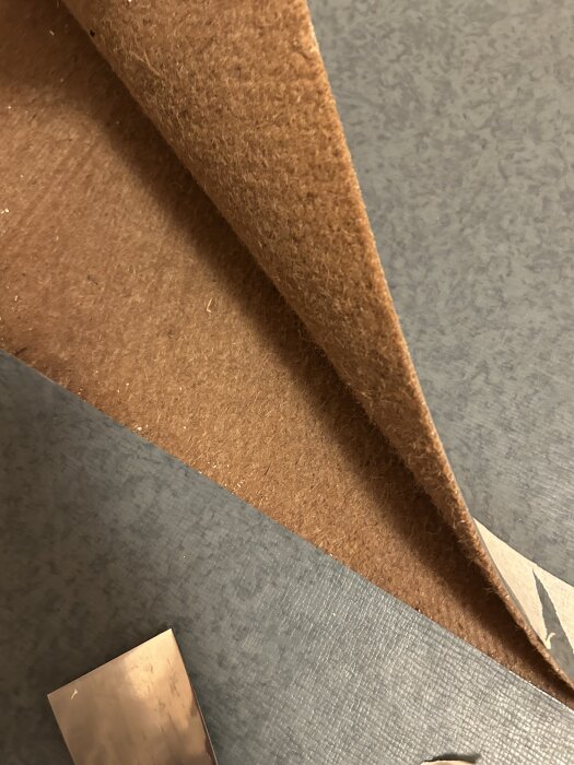 Brun matta från 1966 lyfts för att inspektera om den innehåller asbest, med en suddig bakgrund av golvyta och ett verktyg i hörnet.