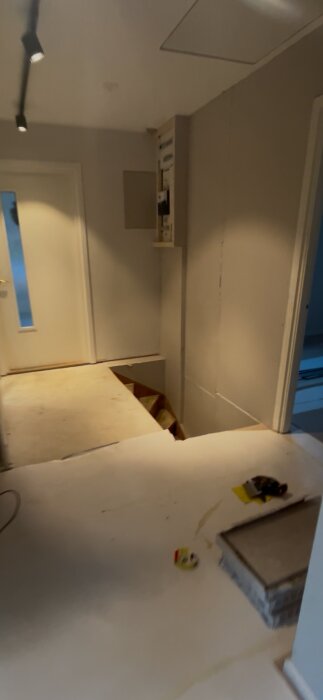 Renovering i en hall med ospacklat golv och avsaknad av trappräcke. Väggarna är vita och det finns en dörr och hängande takbelysning.