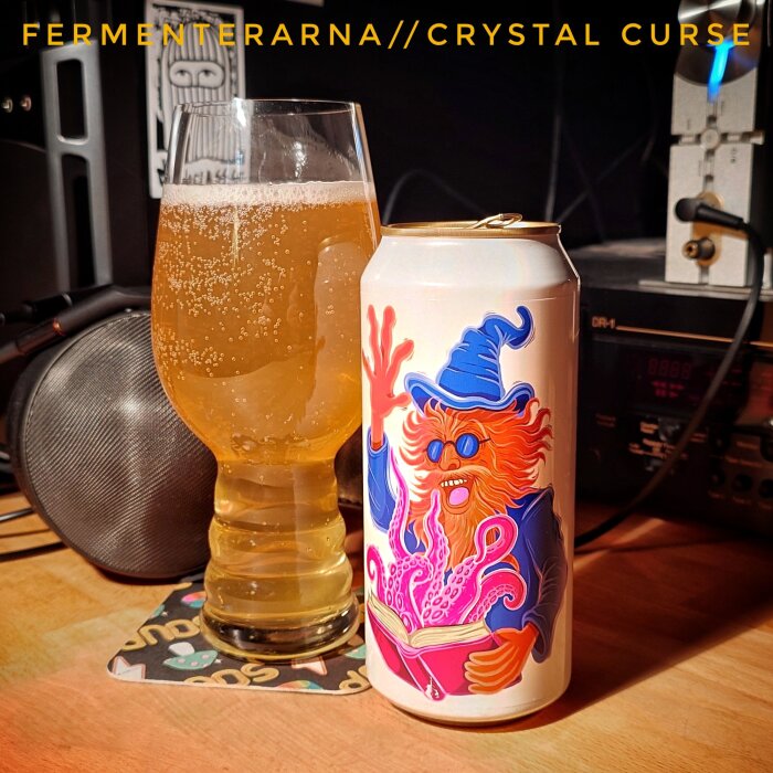 Dryckesglas och burk med färgstark illustration av en trollkarl. Märkt "Fermenterarna//Crystal Curse". Bakgrund med stereoanläggning.