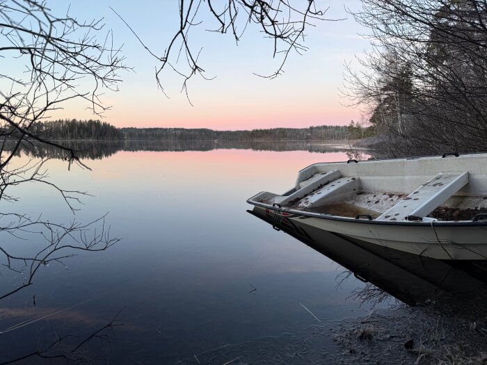 Båt vid isig sjökant med spegelblank vattenyta och skog i bakgrunden, tagen vid solnedgång i vintrig miljö.