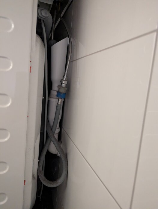Grå slangar och rör bakom en vit panel i ett trångt utrymme.