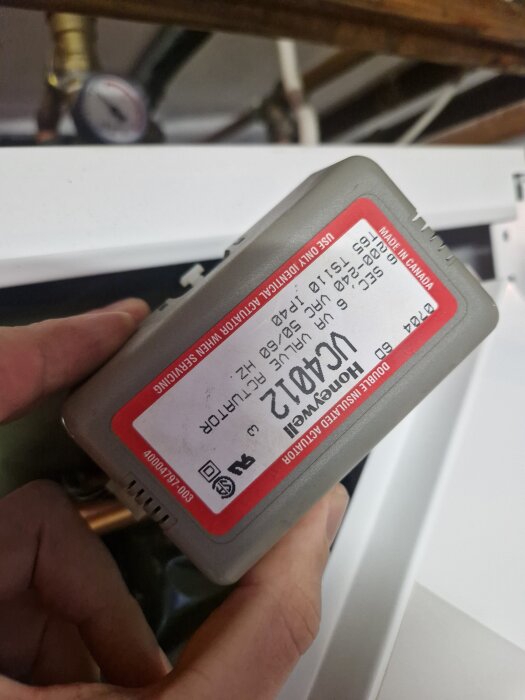 En person håller en Honeywell kompressor med en röd och vit etikett, text och symboler synliga, bakgrunden har ett rör och en tryckmätare.