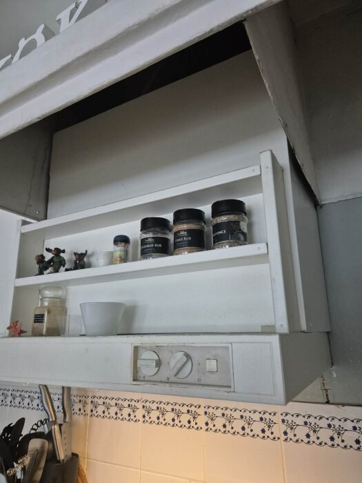 Köksfläkt och förvaringshylla med kryddburkar och dekorativa figurer ovanför spis med vitt kakel och blått mönster.