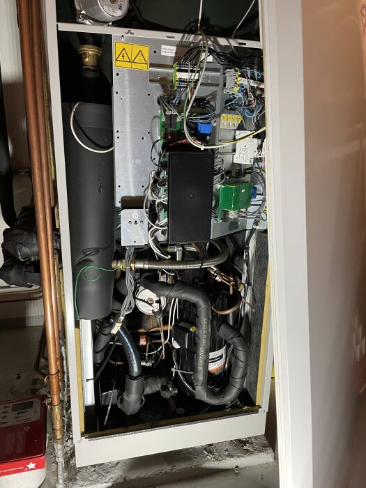 Öppen panel av en värmepump med synliga elektriska komponenter och rörledningar, samt varningsskyltar för högspänning.