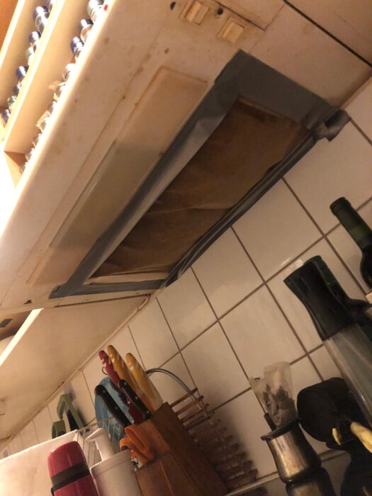Köksfläkt med tejpad ventil och köksredskap på bänken under, i äldre köksmiljö.