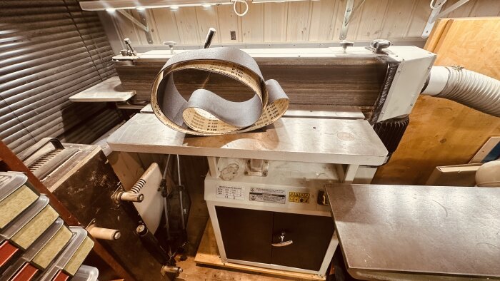Oscillerande bandslipmaskin i ett snickeri med extra slipband P120 placerade på maskinens bord.