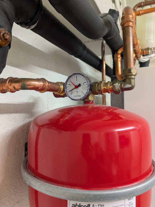 Rött expansionskärl med anslutna kopparrör och en manometer som visar trycket. Svarta isolerade rör syns också i bakgrunden.