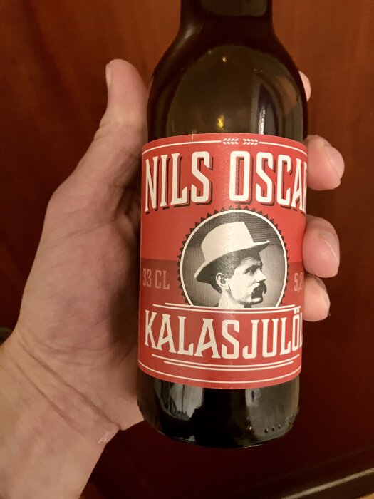 Hand som håller en flaska Nils Oscar Kalasjulöl med rött etikettmotiv av en man med hatt och mustasch, flaskan innehåller 33 cl.