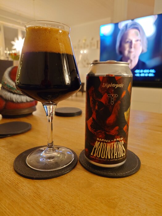 Ett glas med mörk öl och en burk märkt "Barrel-Aged Mountains" på ett träbord, med TV i bakgrunden.