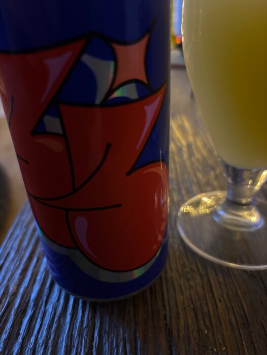 Blå och röd burk med abstrakt mönster och ett glas med gul dryck på ett träbord.