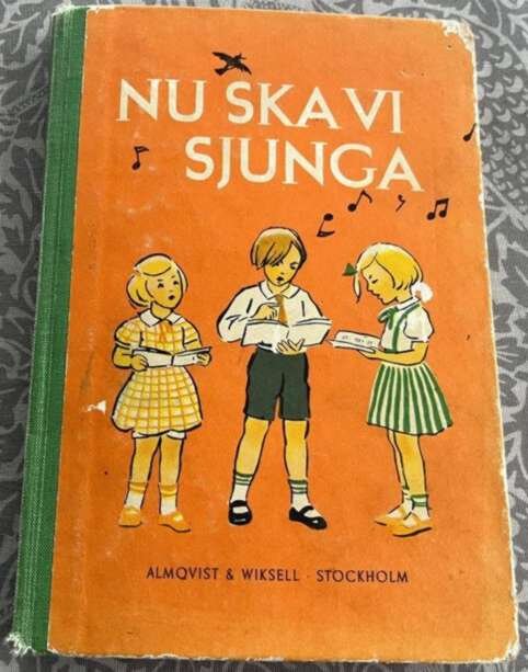 En gammal sångbok med titeln "Nu ska vi sjunga" från 1953, med illustrationer av tre sjungande barn på orange omslag.