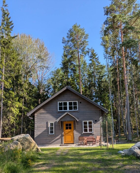 Grått trähus med tjärvitriol, orange dörr och vita fönster i skogsmiljö. Solkatter syns på fasaden.