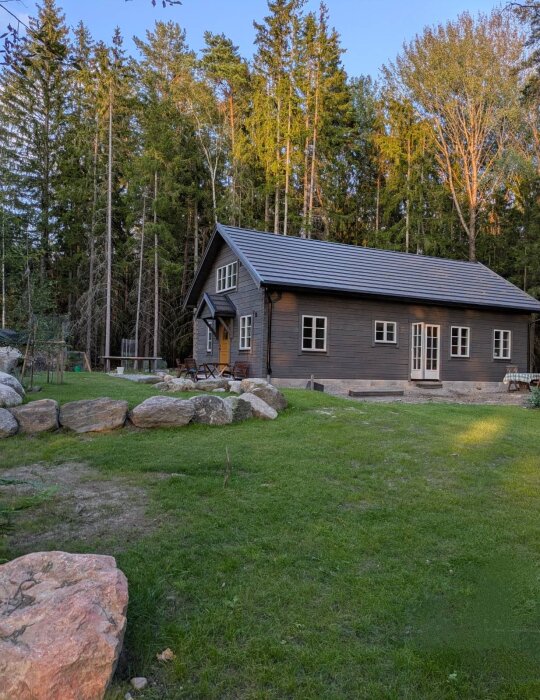 Ett hus beläget på en grön gräsmatta omgivet av stenar, med en skog i bakgrunden och fasad behandlad med tjärvitriol.