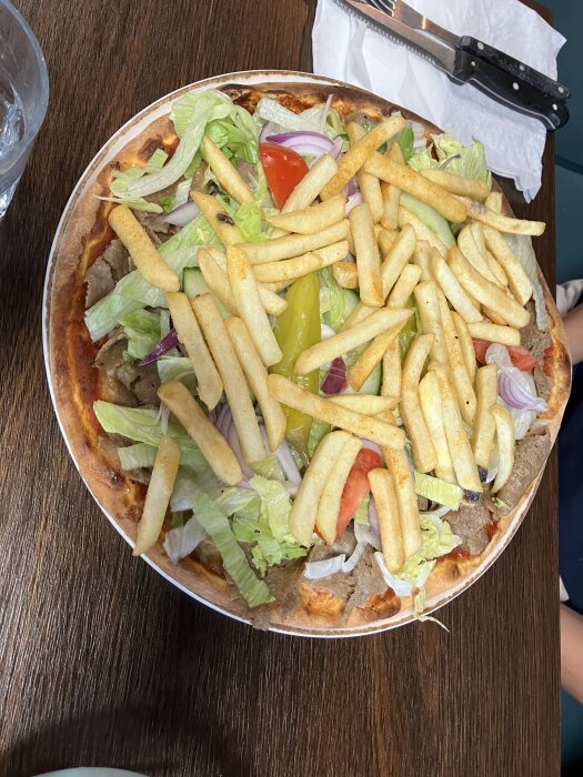 Pizza toppad med pommes frites, kebabkött, sallad, tomat och lök på ett träbord.
