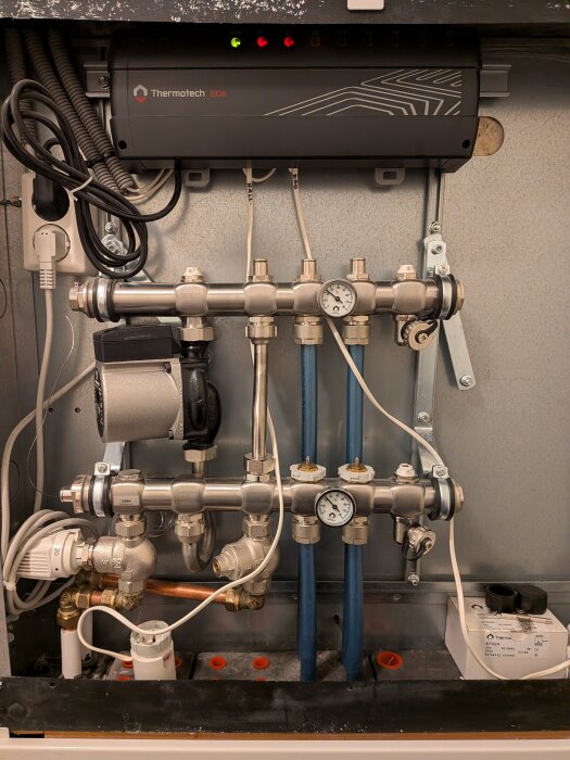Golvvärmeinstallation med kopplingar, rör och två tryckmätare. En Thermotech EC8 enhet syns överst. Kablar och ledningar är anslutna.