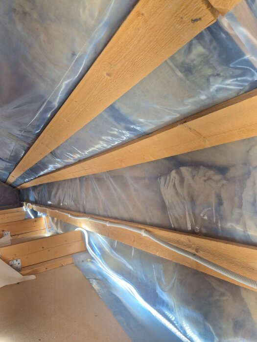Isolerad kattvind med träreglar och ångspärr av plast placerad längs yttertaket, med synlig ventilationsrör.