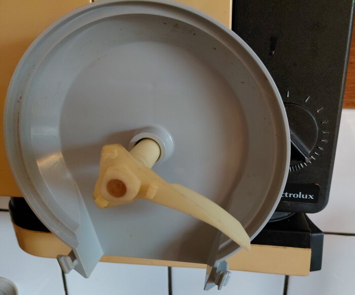 Electrolux köksmaskin med trasig plastvinge, närbild på vit och beige del, mot en brun bakgrund.