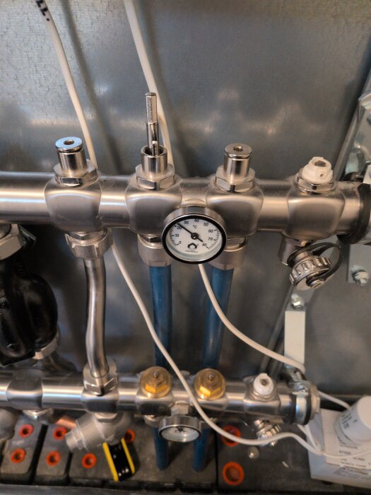 Rörsystem med manometer på ett värmesystem, LED-belysning och kablar synliga. Silvermetalldelar och blå rör är anslutna.