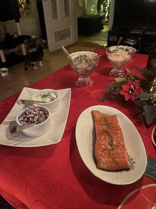 Julbord med lax, gräddfil med dill, hackad lök och två skålar med sillröra på röd duk. Bakgrund med juldekorationer och en katt.