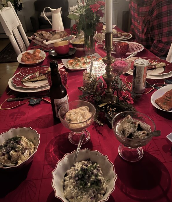 Julbord med olika rätter, inklusive sill och röror, på ett dukat bord med röd duk och blommor som dekoration.