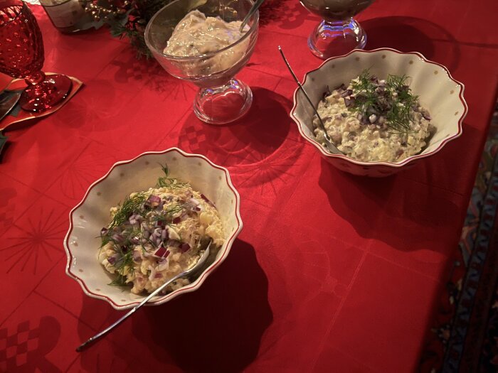 Två skålar med maträtter på ett rött dukat julbord, dekorerade med dill och rödlök, med en tredje skål i bakgrunden.