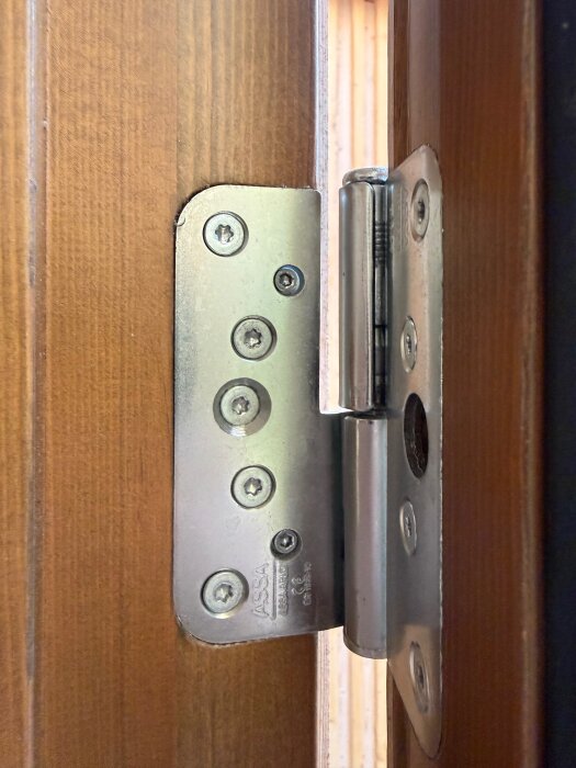 Gångjärn från Assa Abloy monterat på en trädörrkarm, med fem synliga skruvar och metallisk ytfinish.
