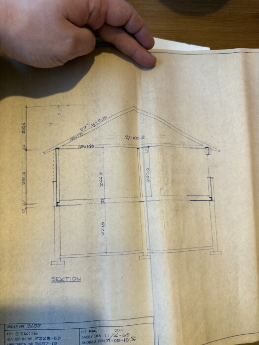 Byggnadsritning för ett hus från 1970-talet, visar sektion med takstolar och källarväggar, inkl. anteckningar och måttangivelser, hålls av en hand.