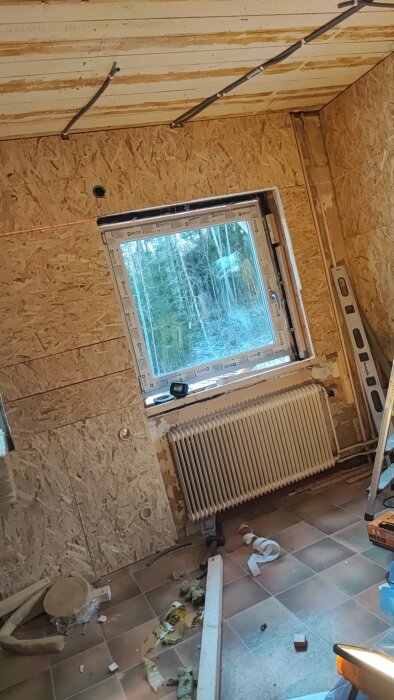 Rum under renovering med nytt fönster installerat, synliga sladdar och verktyg på golvet, samt en stege och nivåverktyg lutade mot väggen.