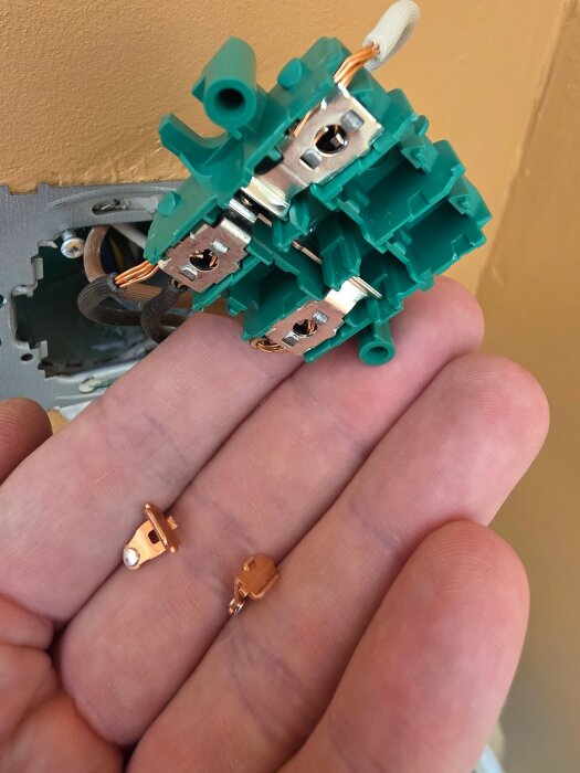 Elektrisk komponent i handen, med kopparkontakter och gröna plastdelar i bakgrunden.