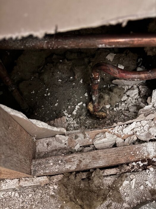Bild av ett hål i en betongvägg med synliga kopparrör, omgiven av byggdamm och isolering i ett pågående renoveringsprojekt.