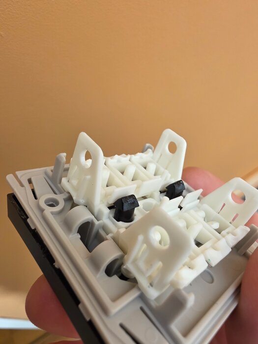 Närbild av vit plastkomponent med clips-fästen, delvis monterad på grå bas, hålls i en hand mot en beige bakgrund.